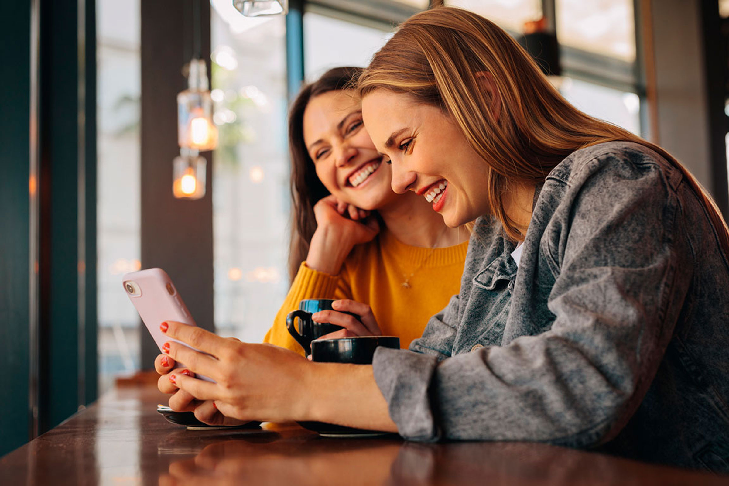 Zwei junge Erwachsene sitzen lächelnd auf einer Couch und schauen gemeinsam auf ein Handy. Das Bild vermittelt eine entspannte Atmosphäre bei der Erledigung von Bankgeschäften und verweist auf attraktive BBVA Aktionen wie Cashback oder das kostenlose Girokonto.