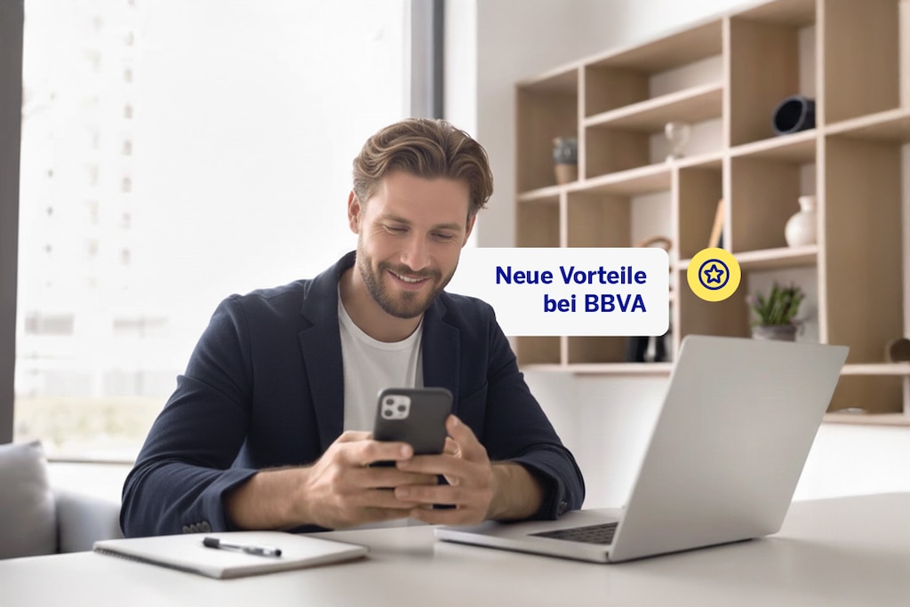 Eine Person sitzt am Schreibtisch vor einem Laptop und liest auf ihrem Handy die Vorteile des Gehaltskontos bei BBVA.