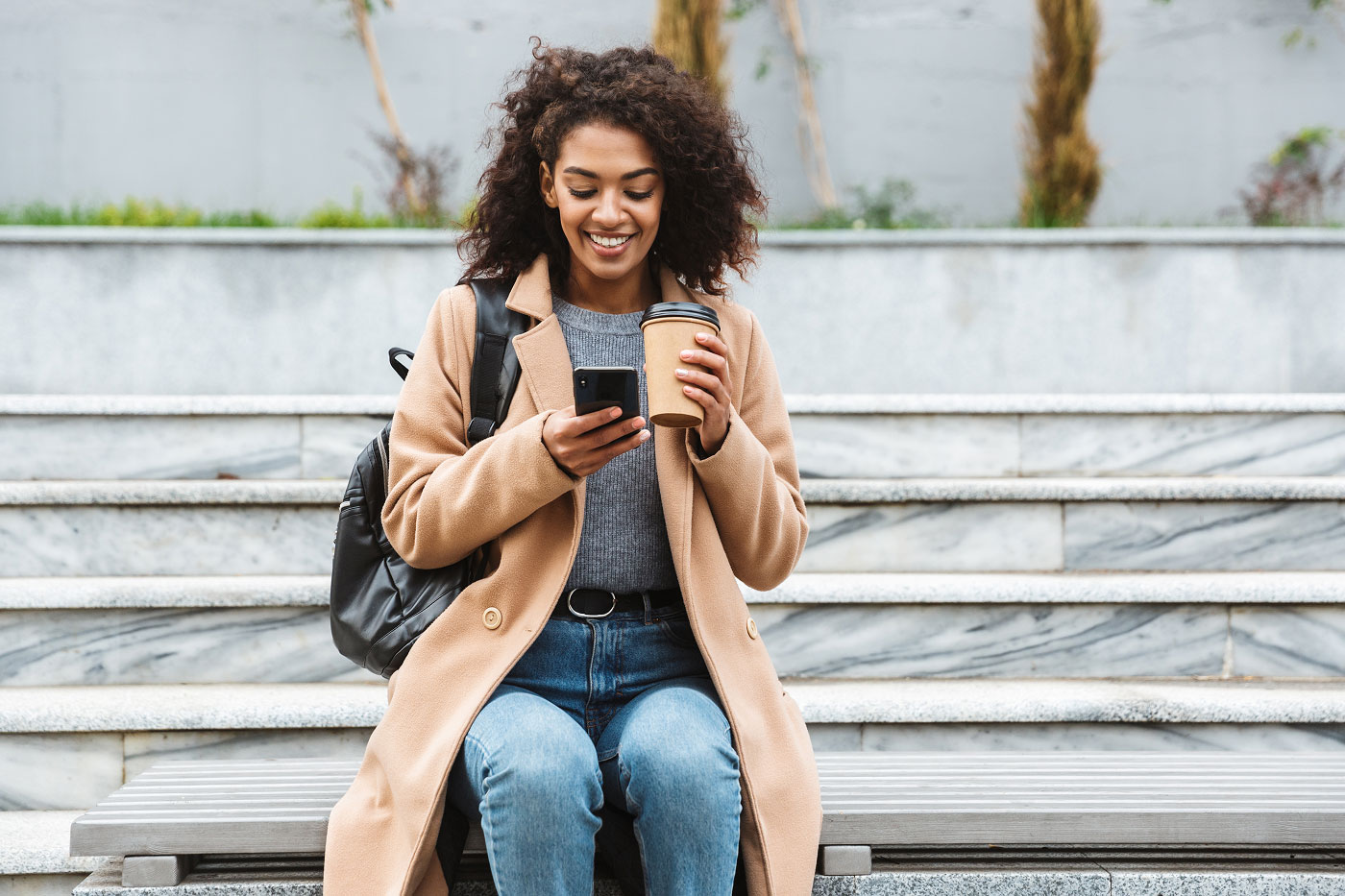 A woman with black curls and a backpack is sitting on an outdoor staircase and using her smartphone to manage her debit card.