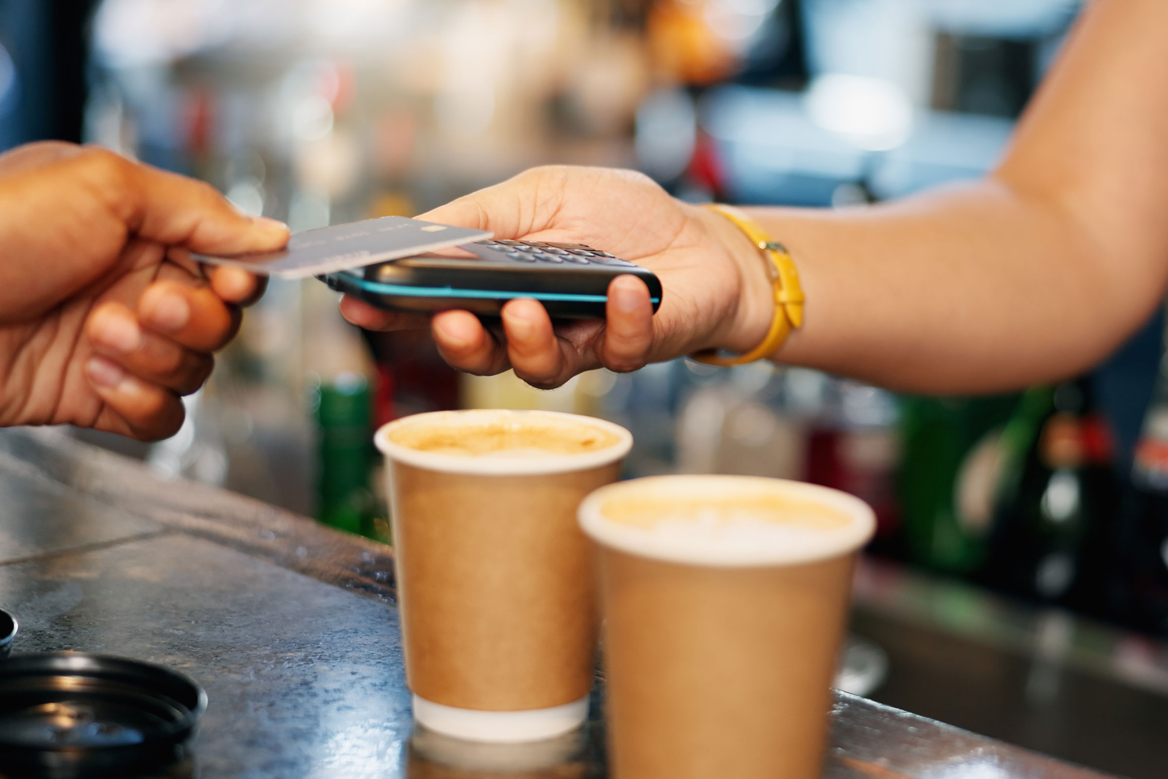Close-up of hands at a counter paying for two cups of coffee using a contactless debit card at a terminal.